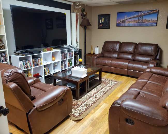 A living room features two brown leather couches facing a coffee table, with a large television mounted on the wall and a bookshelf filled with DVDs in the corner.