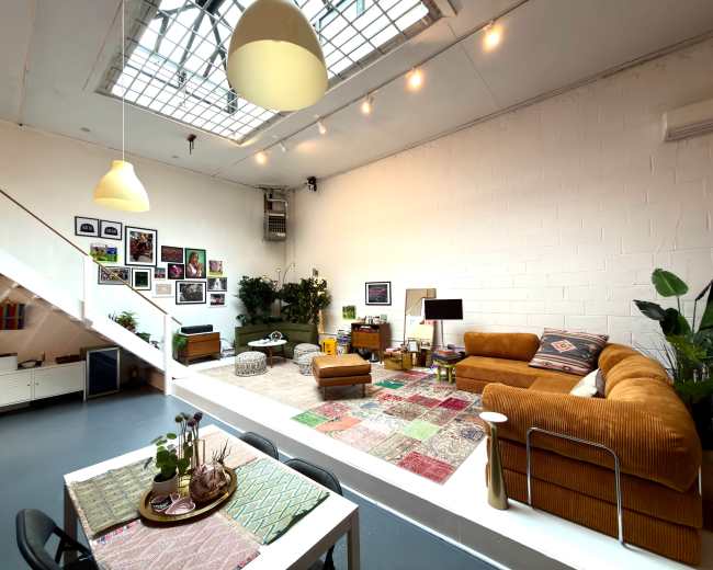 The image shows a spacious, well-lit living room featuring a mix of furniture, plants, and artwork.