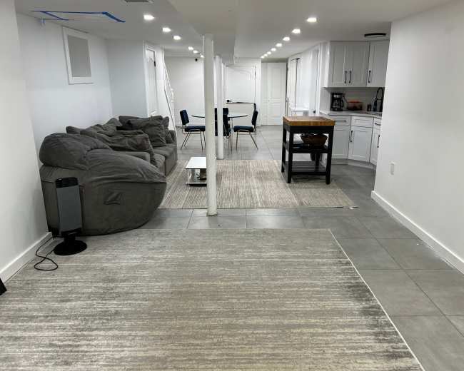 The image shows a spacious basement area featuring a gray sectional couch, a dining table with chairs, and a small kitchen island.