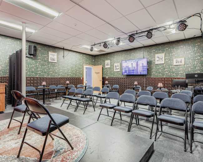 The image shows a small auditorium with rows of blue chairs facing a wall-mounted television and a stage area, set against a patterned green backdrop.
