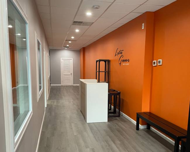 The image shows a narrow hallway with gray walls and an orange accent wall, featuring a reception area with a white counter and dark seating along one side.