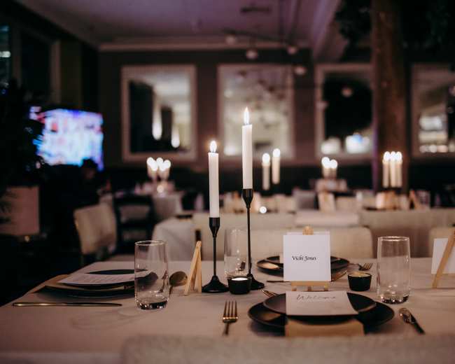 The image shows a neatly arranged dining area with tables set for an event, featuring candles, place cards, and glassware.