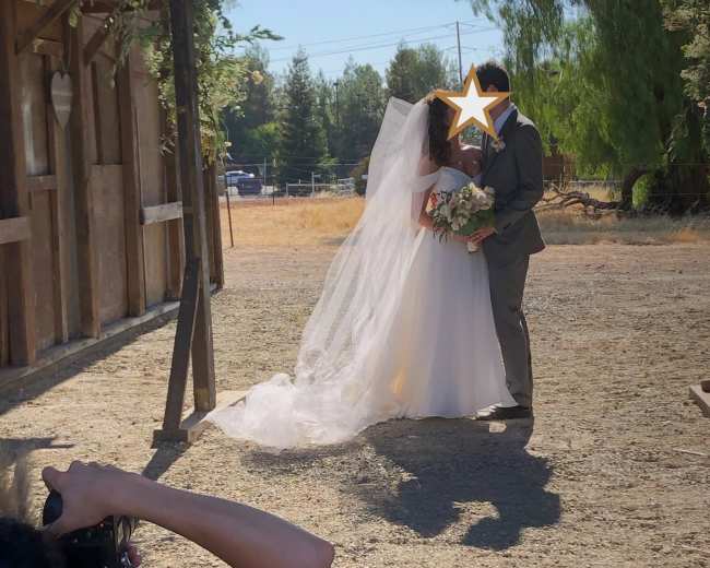 A bride and groom embrace in a sunlit outdoor setting, while a photographer captures the moment nearby.