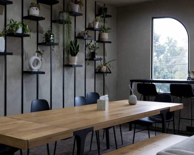 A long wooden table is set in a modern room with plants displaying on wall-mounted shelves and a bar area in the background.
