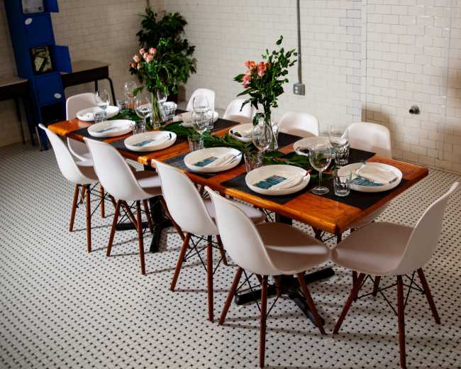 A long wooden dining table is set with plates and glassware, surrounded by white chairs in a tiled room with plants and a blue storage unit.