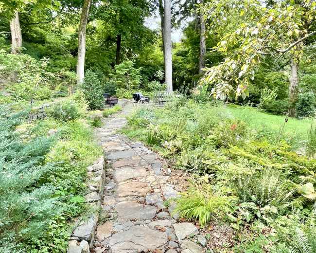 A stone pathway winds through a lush garden surrounded by trees and various plants.