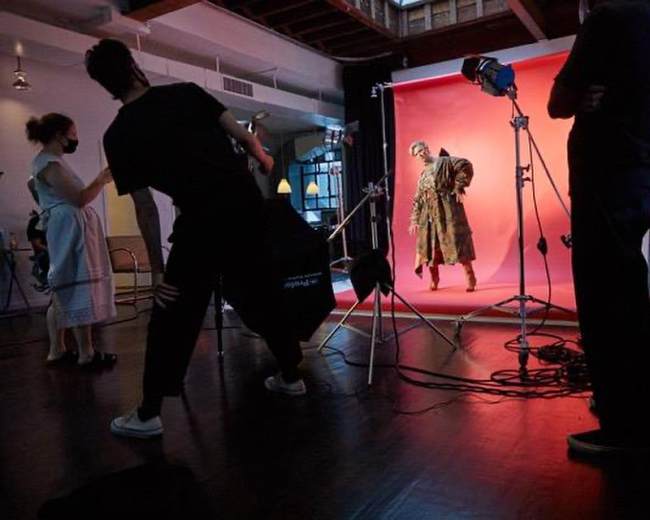 A photoshoot is taking place in a studio with a model posing against a pink backdrop while crew members adjust equipment.