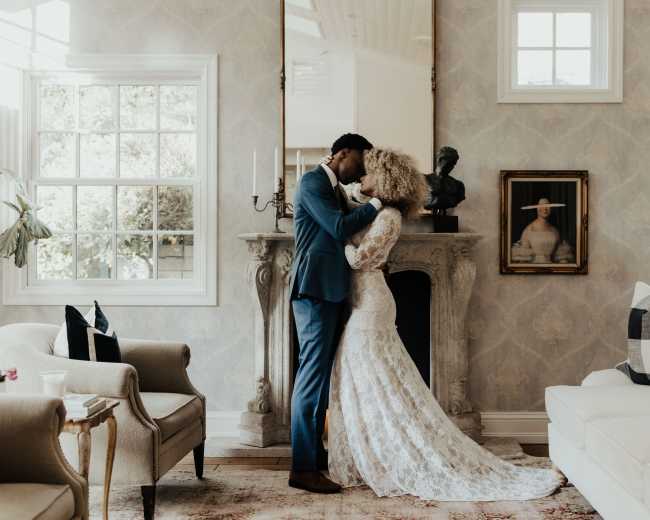 A couple embraces in a stylish living room with a fireplace and large windows.