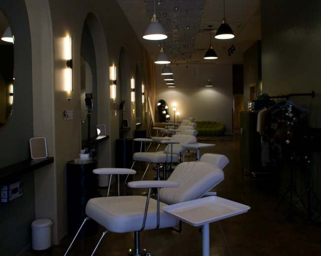 The image shows a modern salon interior with several white treatment chairs lined up along a long corridor, illuminated by overhead pendant lights.