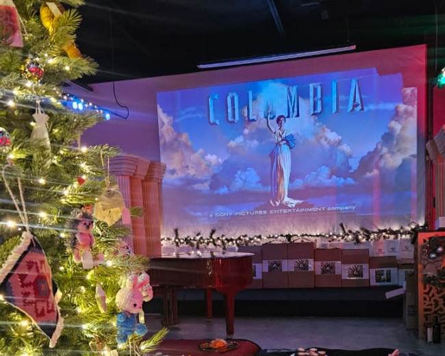 The image shows a cozy indoor scene with a decorated Christmas tree on the left and a projection of "COLUMBIA" on the wall in the background.
