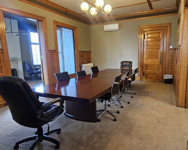 The image shows a conference room with a large wooden table surrounded by black office chairs, adorned with wooden paneling and large windows.