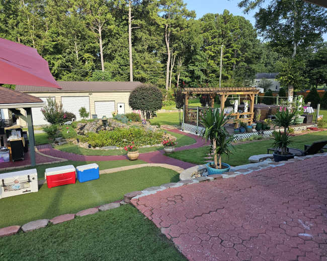 The image shows a landscaped backyard with a patio area, flower beds, garden structures, and a small water feature surrounded by greenery.