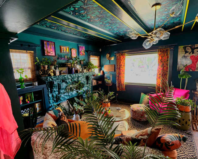 The image shows a vibrant living room filled with plants, colorful furniture, and artistic decorations on the walls.
