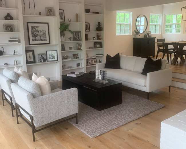 The image shows a living room with a sofa and two armchairs around a coffee table, surrounded by built-in shelves filled with decorative items, and a dining area visible in the background.