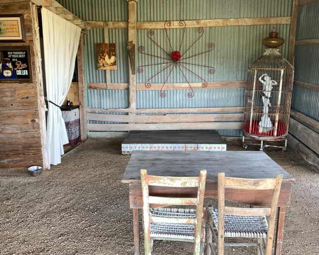 The image shows an interior space with wooden walls, a small table, two wooden chairs, and a decorative birdcage against a backdrop of corrugated metal.