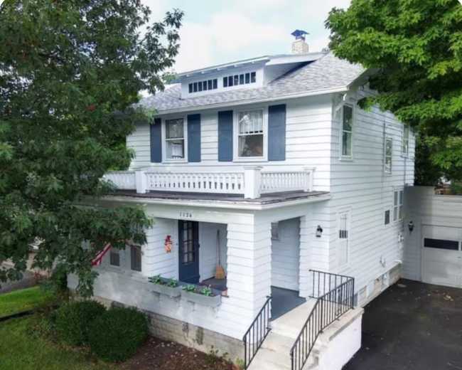 The image shows a two-story white house with blue shutters and a front porch, surrounded by greenery and parking space.