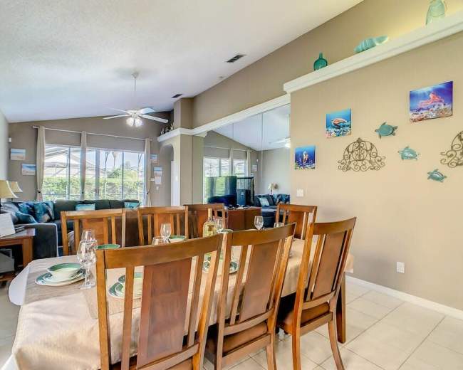 The image shows a dining area with wooden chairs set around a table, adjacent to a living space featuring a large couch and a TV in the background.