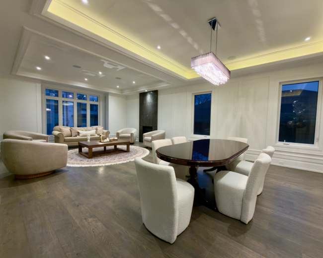 The image shows a modern, elegantly designed living space featuring a dining table surrounded by plush chairs, a cozy seating area with a sofa, and large windows allowing natural light to enter.