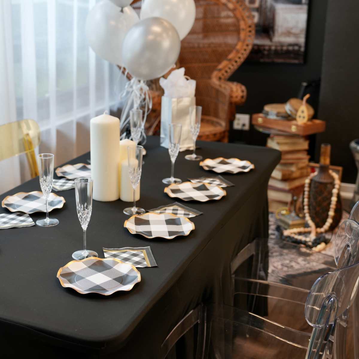 The table is set with black and white checkered plates, champagne flutes, and a lit candle, surrounded by transparent chairs and decorative balloons.
