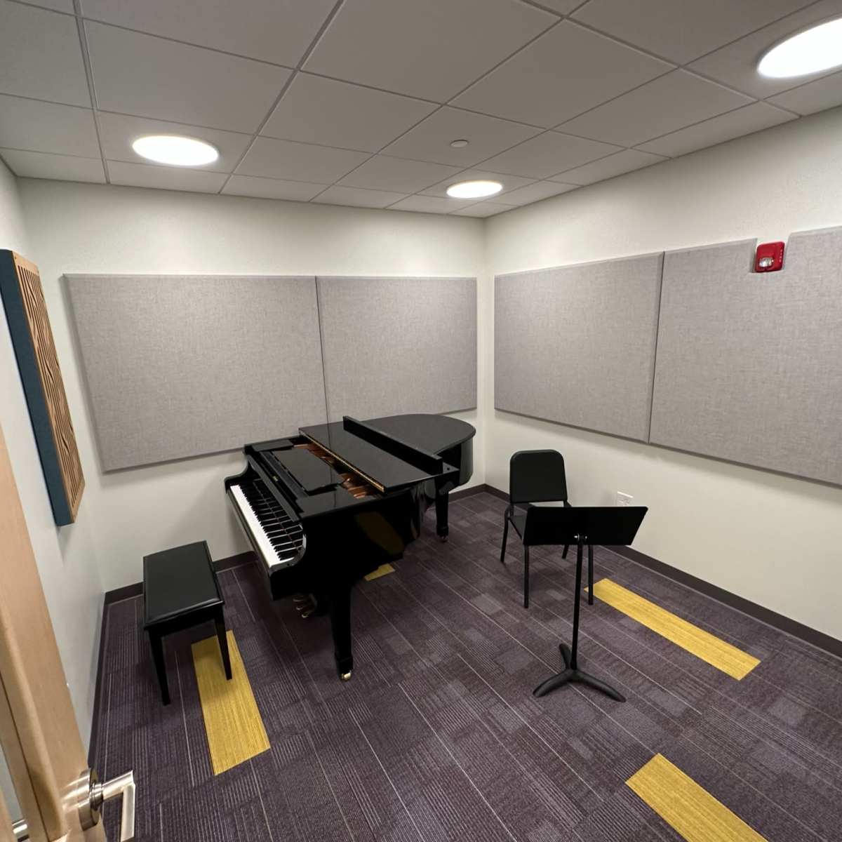 A small music practice room featuring a grand piano, a chair, and a music stand, with sound-absorbing panels on the walls.