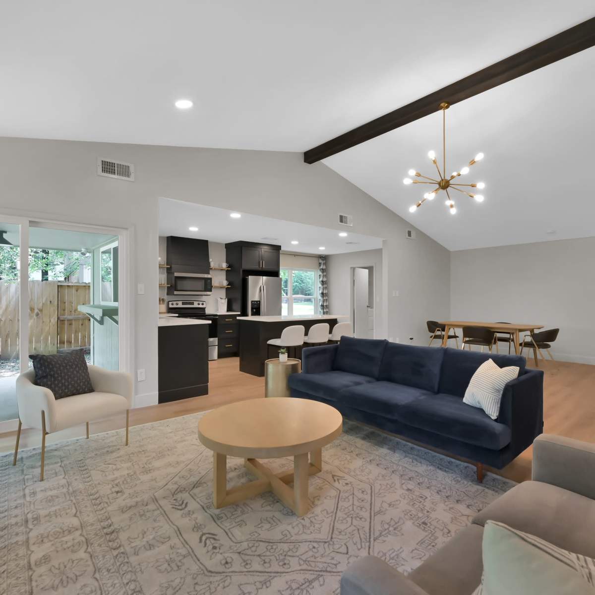 A modern living room with a blue sofa, a round wooden coffee table, and a dining area in the background, featuring a kitchen with black cabinetry.