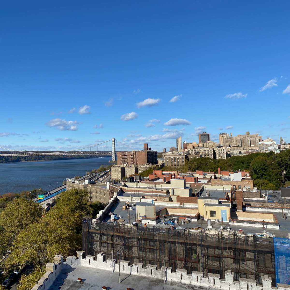 A view of the Hudson River with the George Washington Bridge in the background, and buildings lining the riverbank, including a neighborhood with low-rise structures and trees.