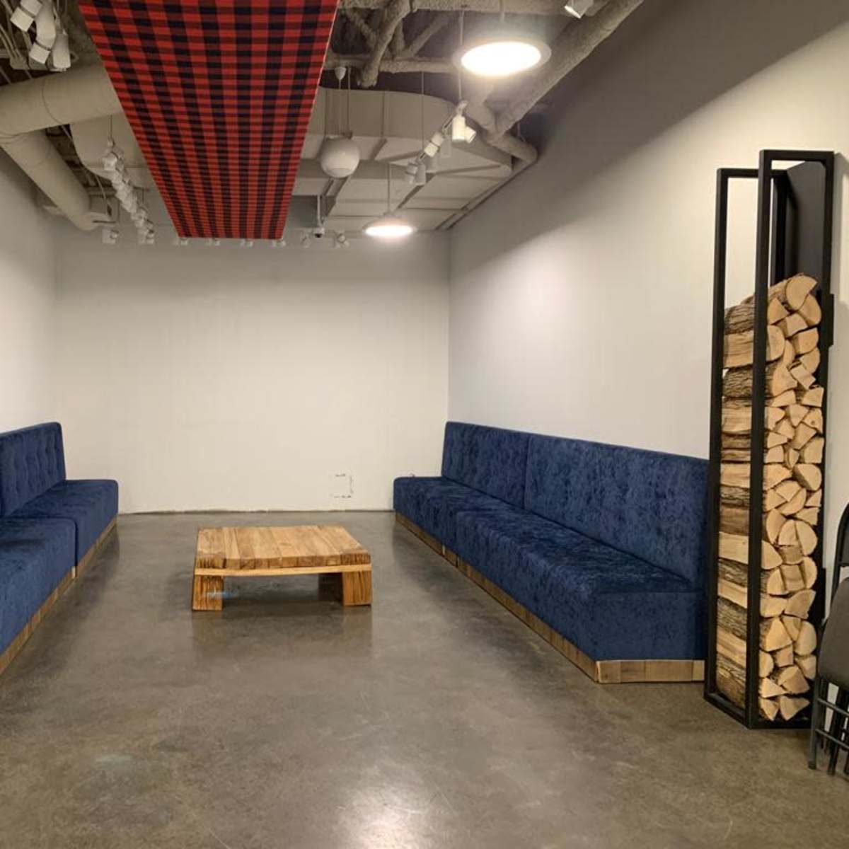 A modern waiting area features two long blue sofas, a wooden coffee table in the center, and decorative wooden logs stacked on either side.