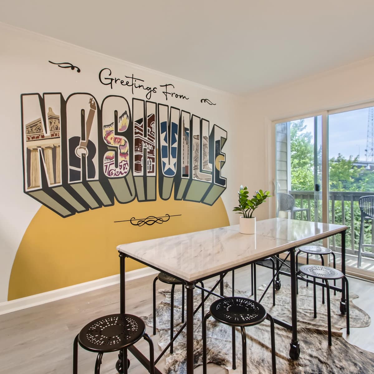 A modern dining area with a large marble table surrounded by black chairs, set against a wall featuring a colorful "Greetings From Nashville" mural and a view of the outdoors through a sliding glass door.