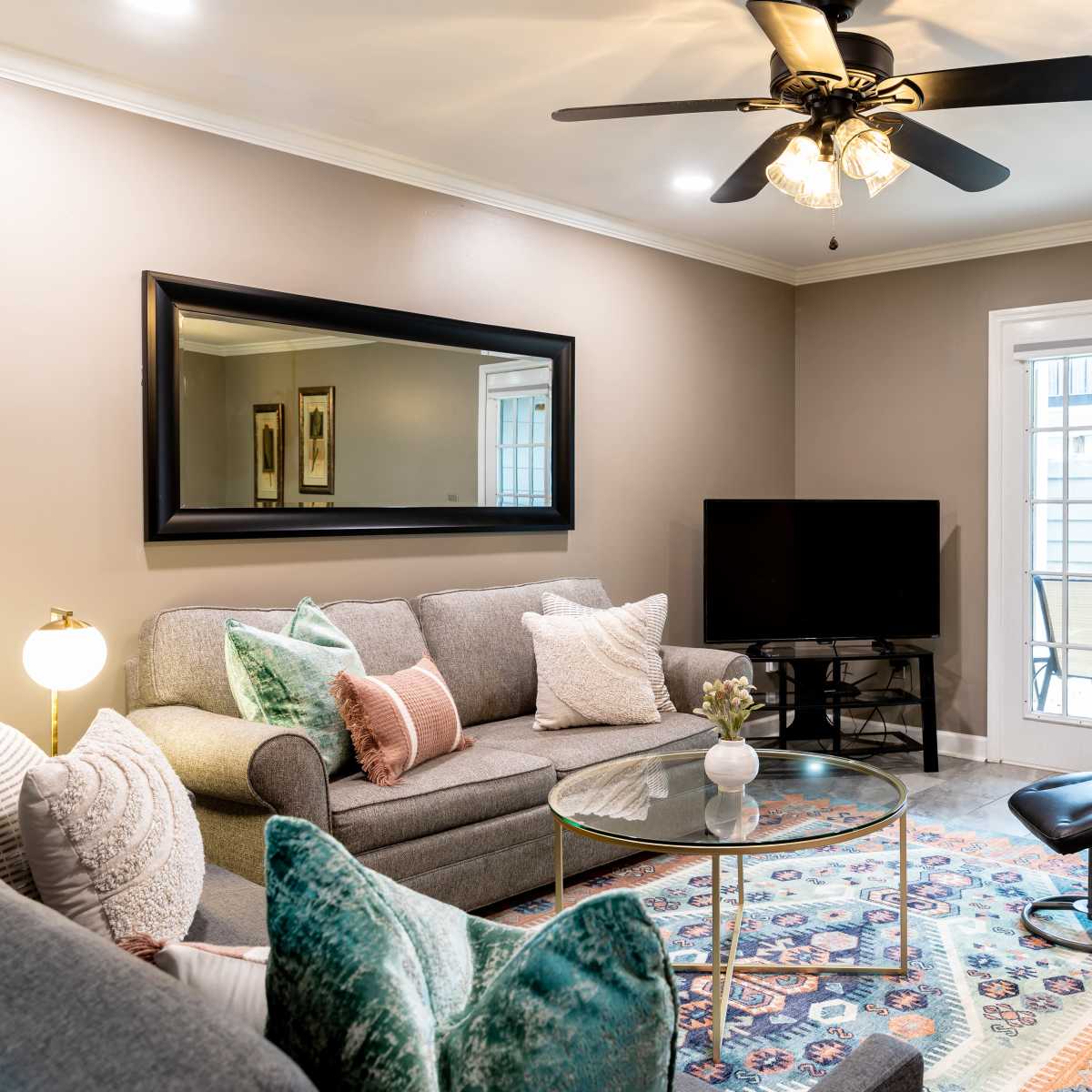A living room featuring a gray couch, a glass coffee table, a television on a stand, and a black reclining chair, with a ceiling fan above and patio doors leading outside.