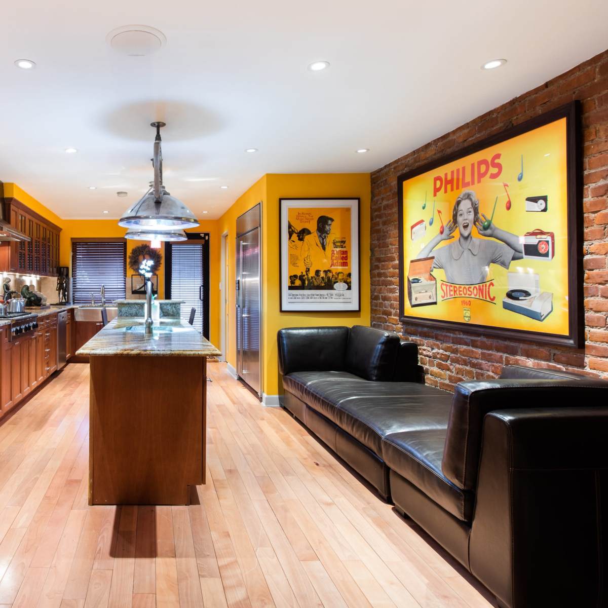 A kitchen with wooden cabinetry, a granite island, and a yellow and red color scheme, featuring a brick wall and vintage posters.