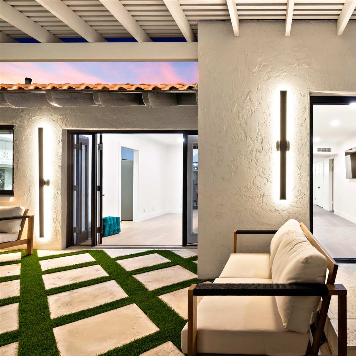 A modern outdoor seating area with a stone patio, green grass accents, and large glass doors leading to an interior space.