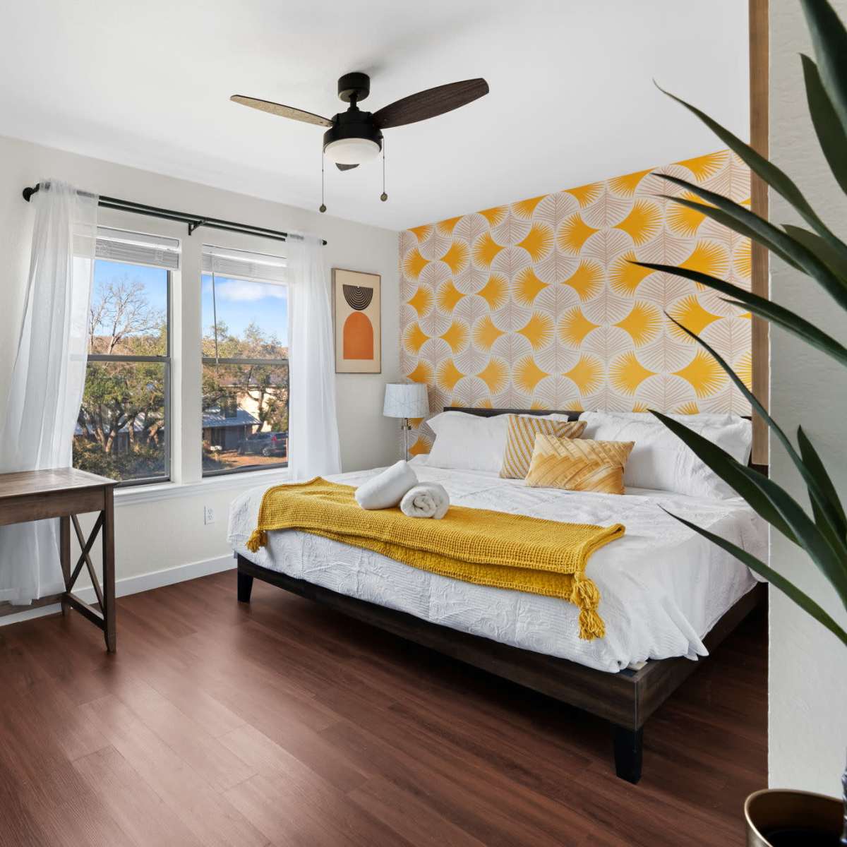 A bright bedroom featuring a bed with patterned yellow and white accents, a desk with a chair, and a window allowing natural light.