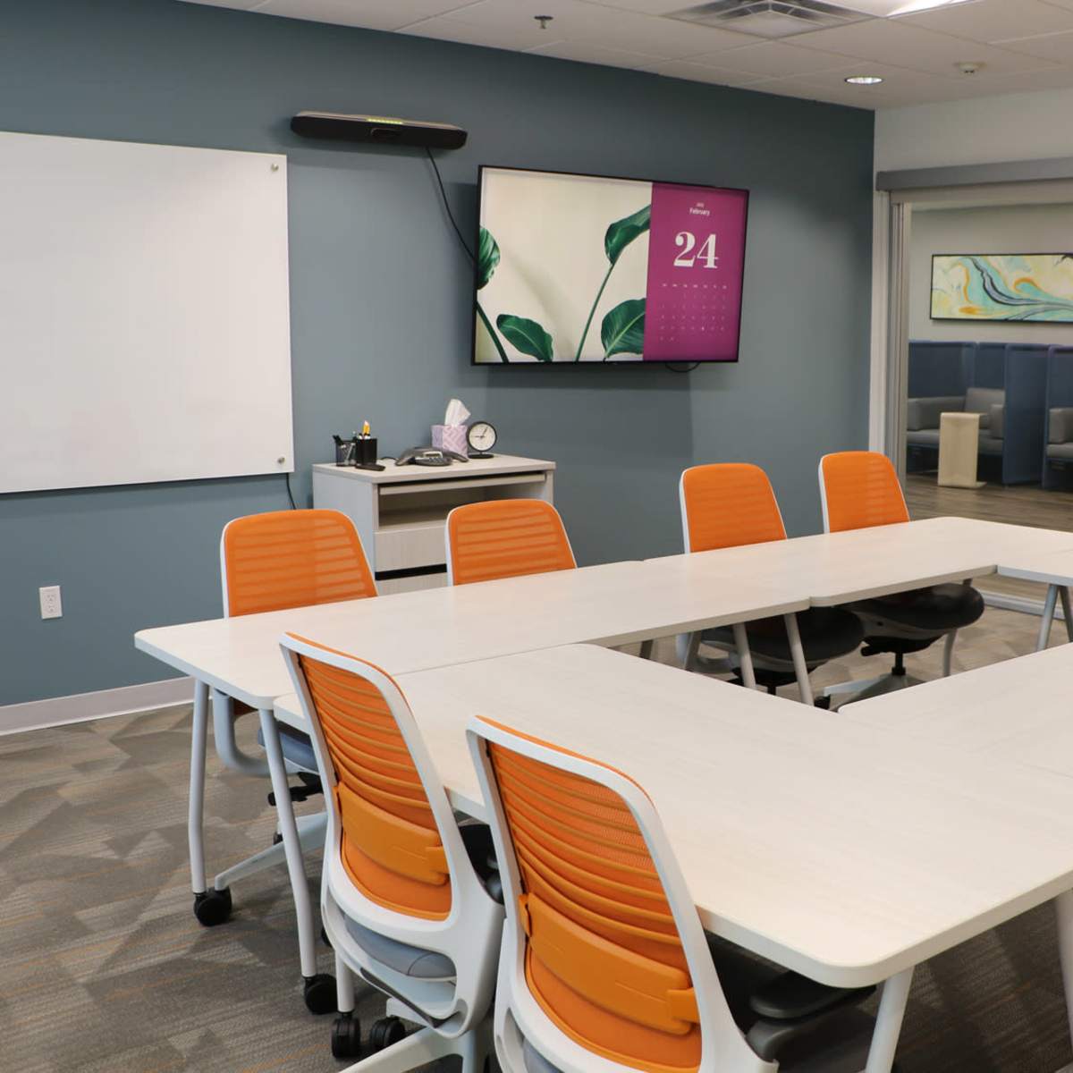 A modern conference room with a large white table arranged in a U-shape, surrounded by orange and white chairs, and equipped with a wall-mounted display and a whiteboard.