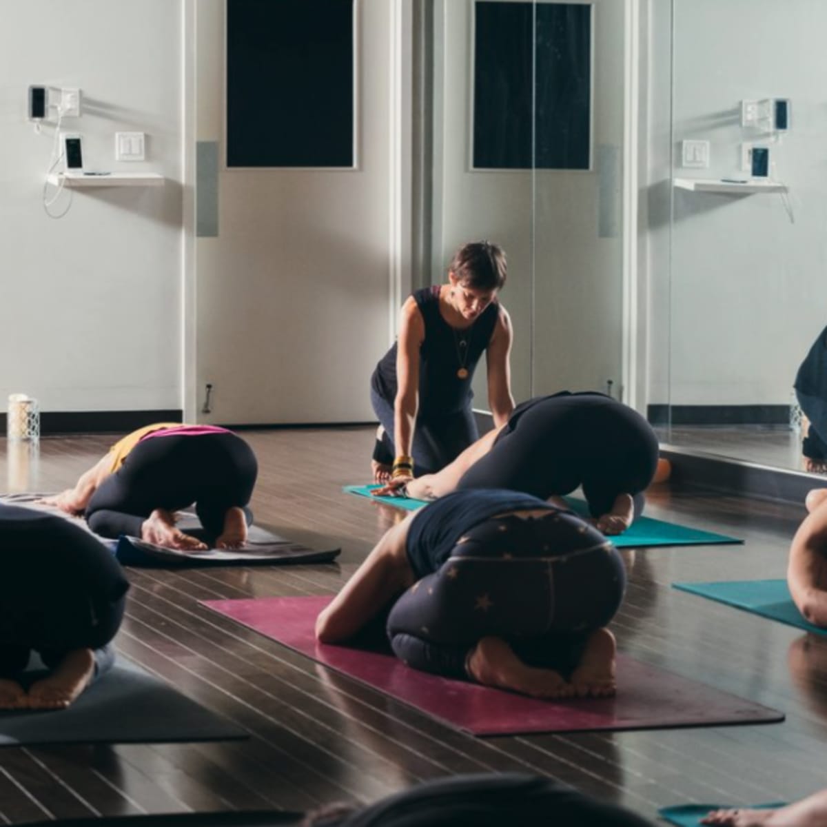A group of individuals practices yoga in a studio, with one instructor assisting a participant while others are in a forward bend position on their mats.