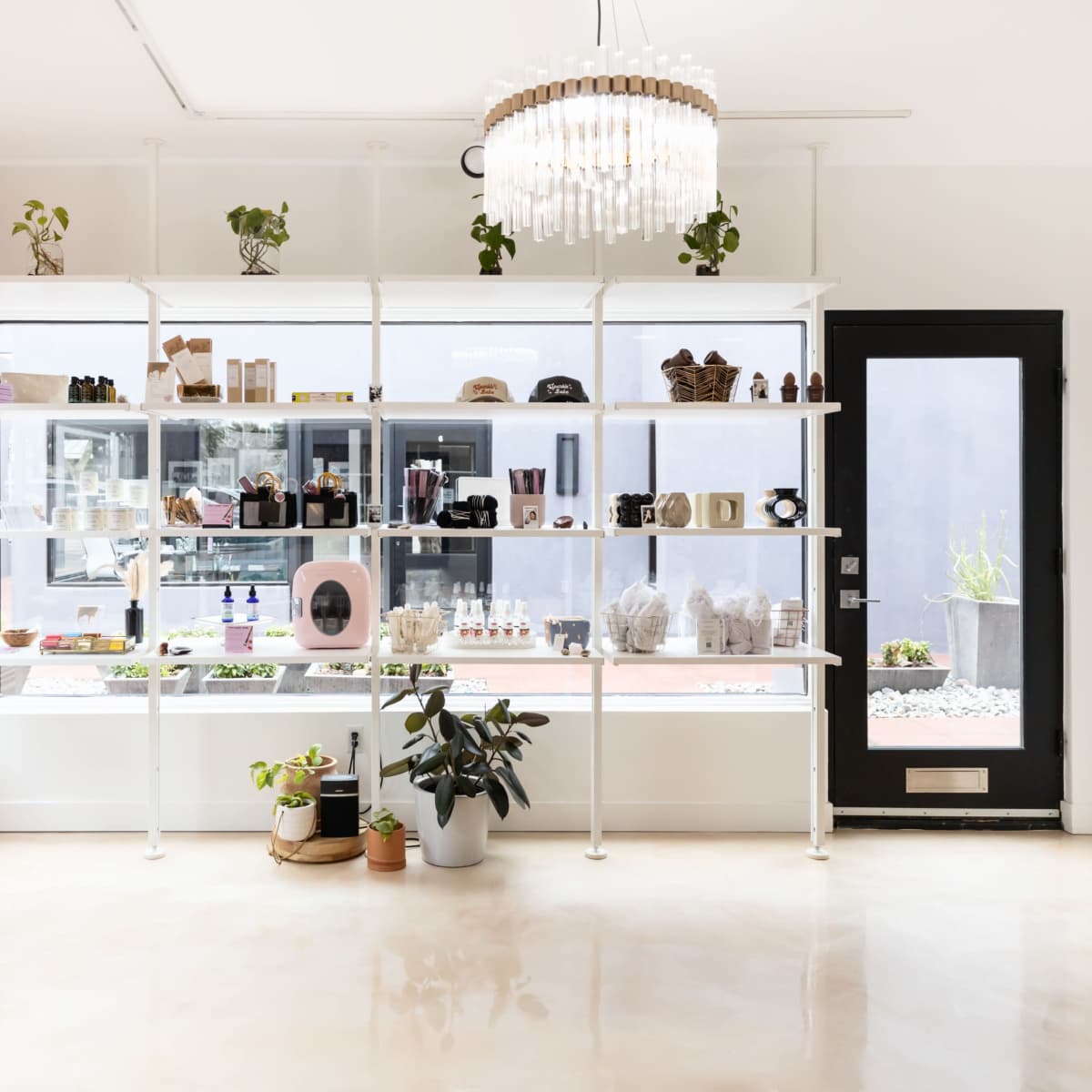 A modern boutique shop with white shelving displaying various products under a chandelier, alongside a large glass door.