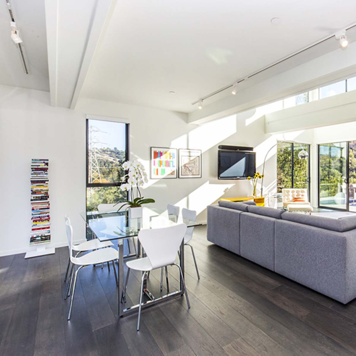 A modern living space featuring a gray sectional sofa, a glass dining table with white chairs, and large windows that provide a view of the surrounding hillside.