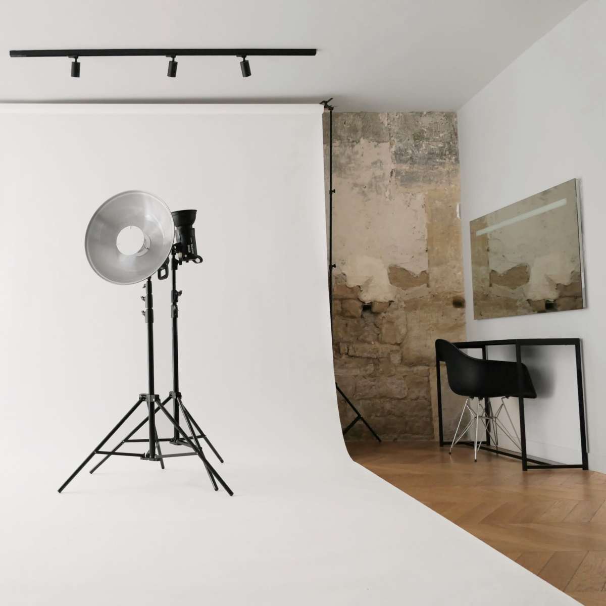A photography studio setup with a white backdrop, a large light fixture on a stand, and a chair beside a mirror against a textured wall.