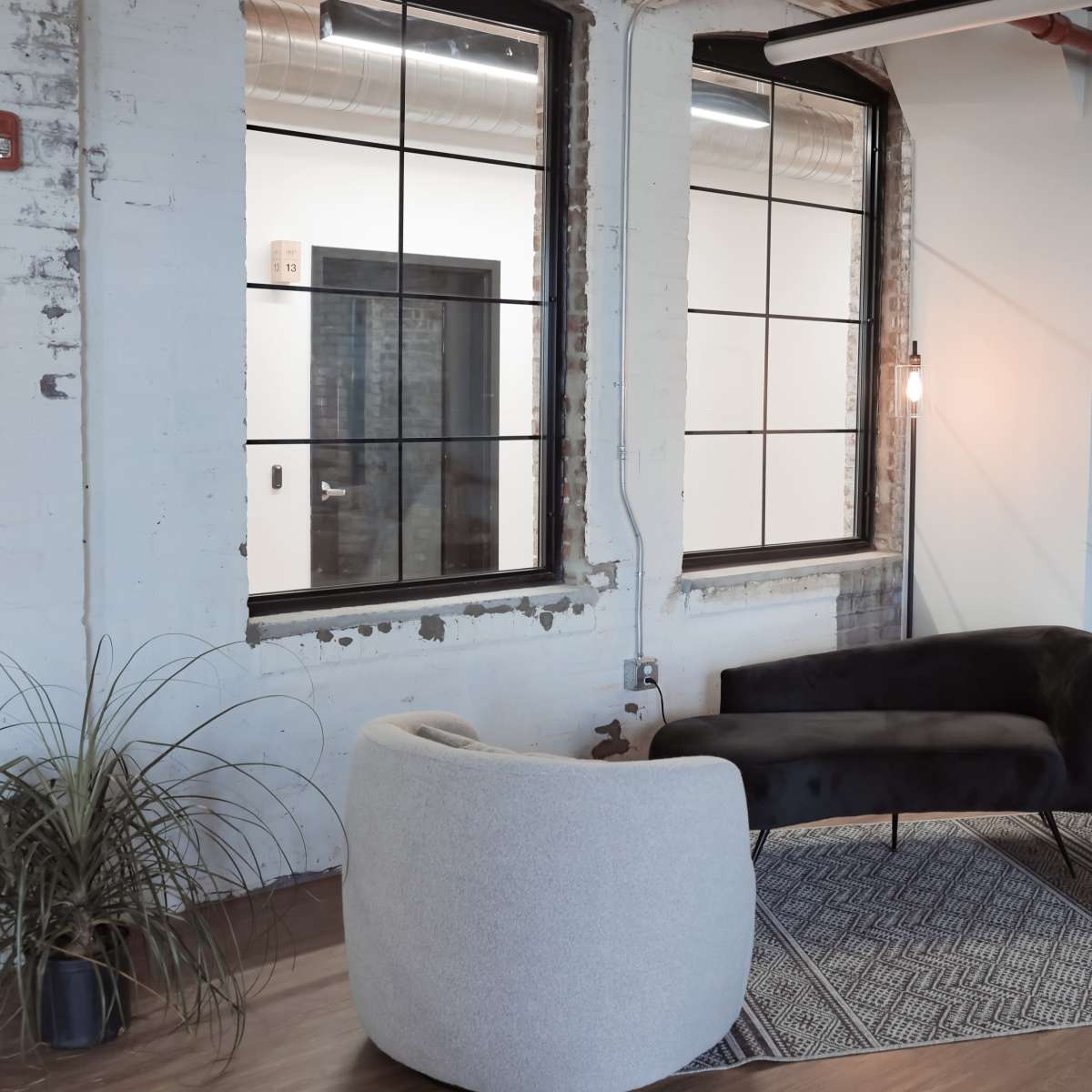 A seating area with two curved armchairs and a black velvet sofa arranged around a patterned rug, positioned near large windows with black frames.