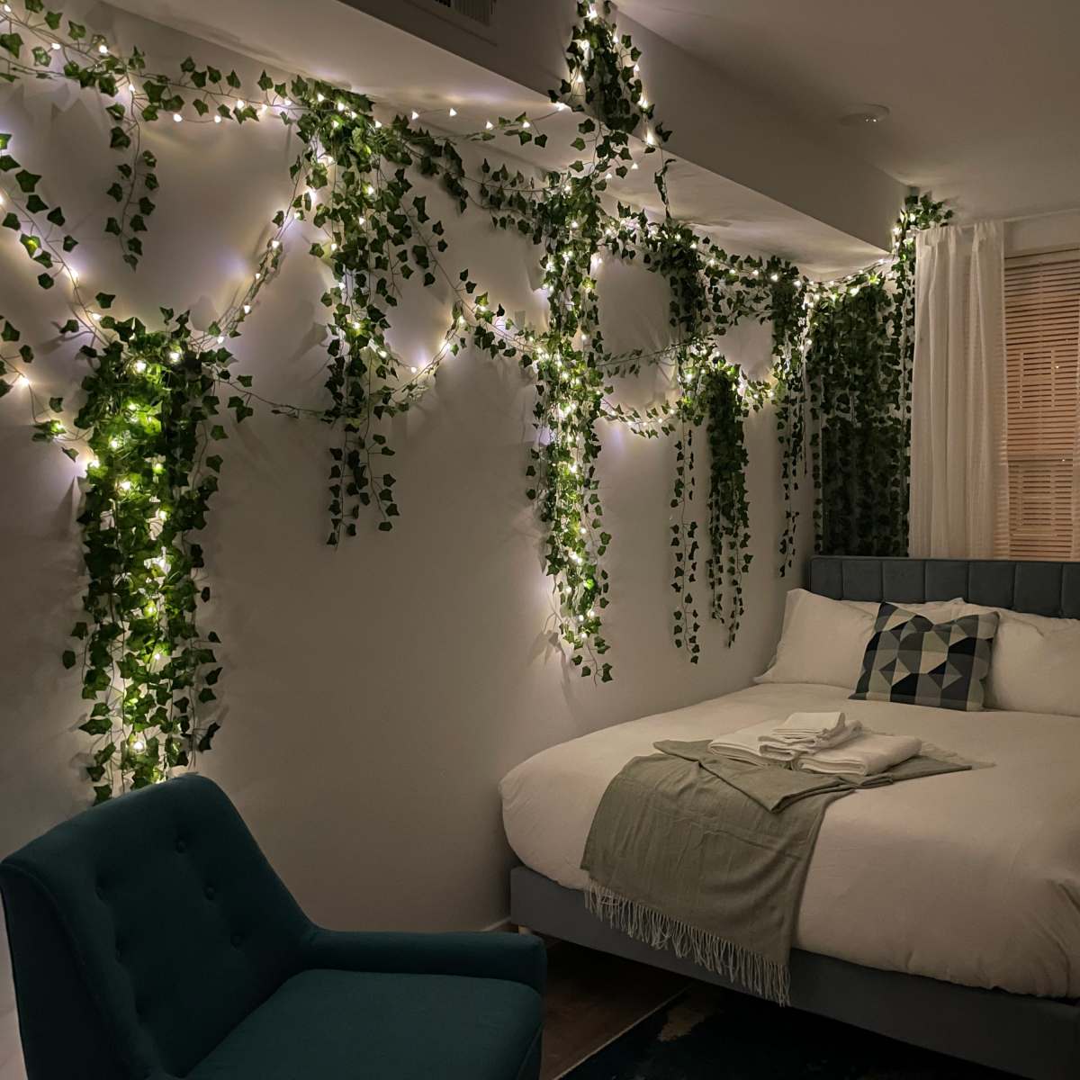 A bedroom with a bed, decorated wall with green vines and string lights, a blue chair, and a window covered with sheer curtains.