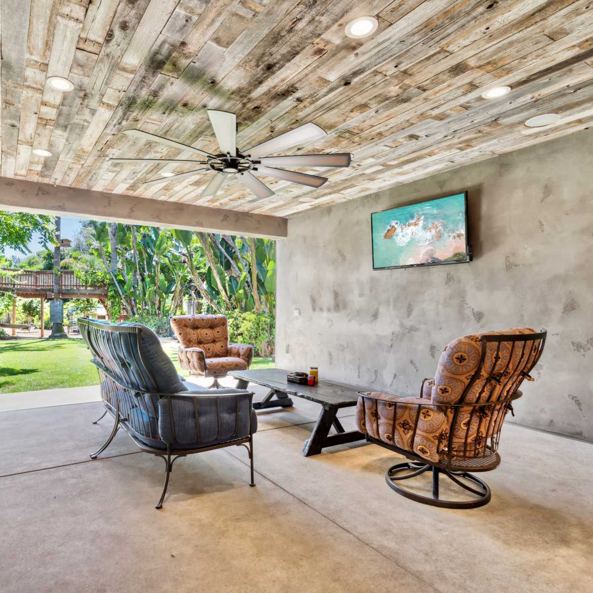 A covered patio features two chairs and a coffee table, overlooking a landscaped yard with tropical vegetation.