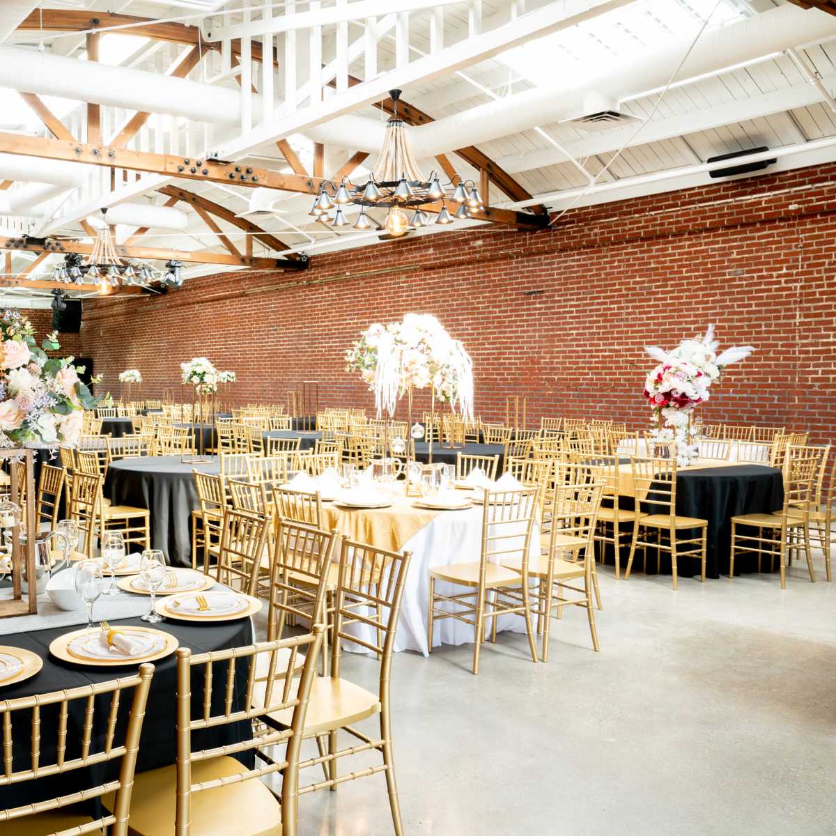 A spacious event hall features elegantly arranged tables with black and white tablecloths, gold chairs, and floral centerpieces against a backdrop of exposed brick and wooden beams.
