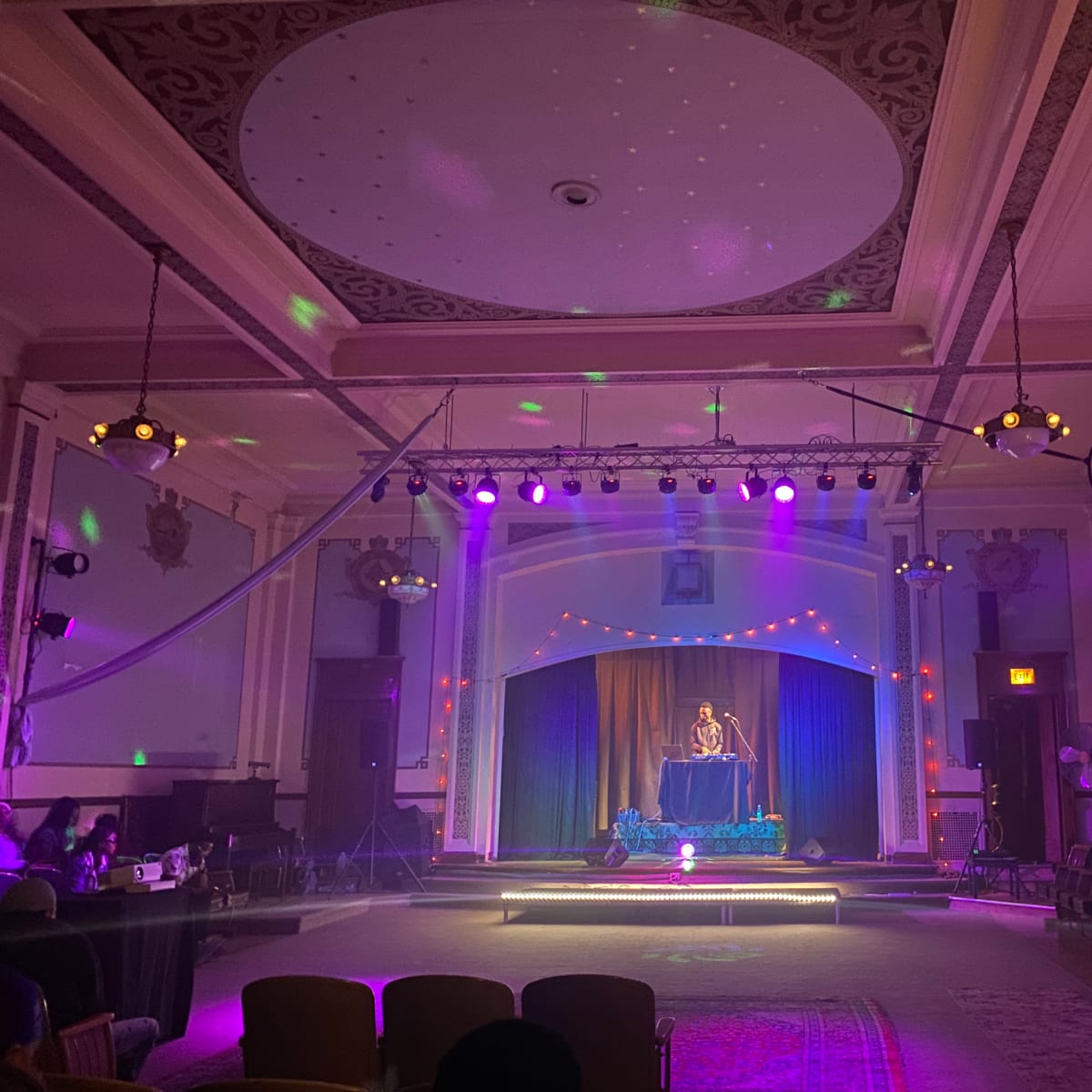 A performer stands on a stage adorned with lights in a decorated hall, surrounded by empty chairs.