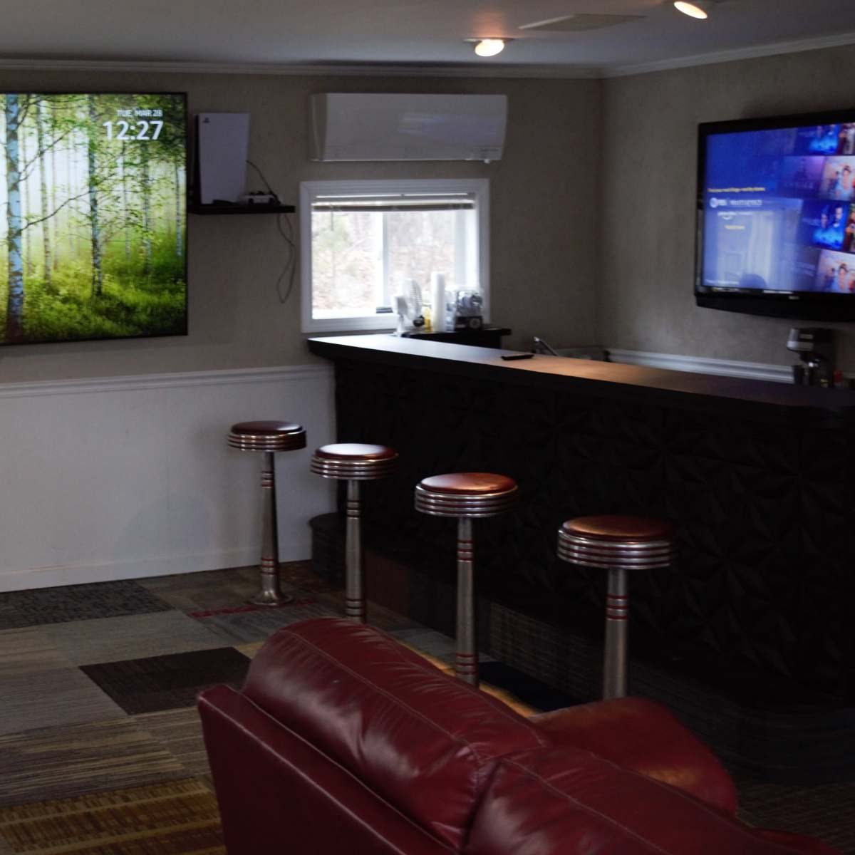 A room with a large TV displaying a forest scene, a bar area with four stools, and a couch in front of a chess table.