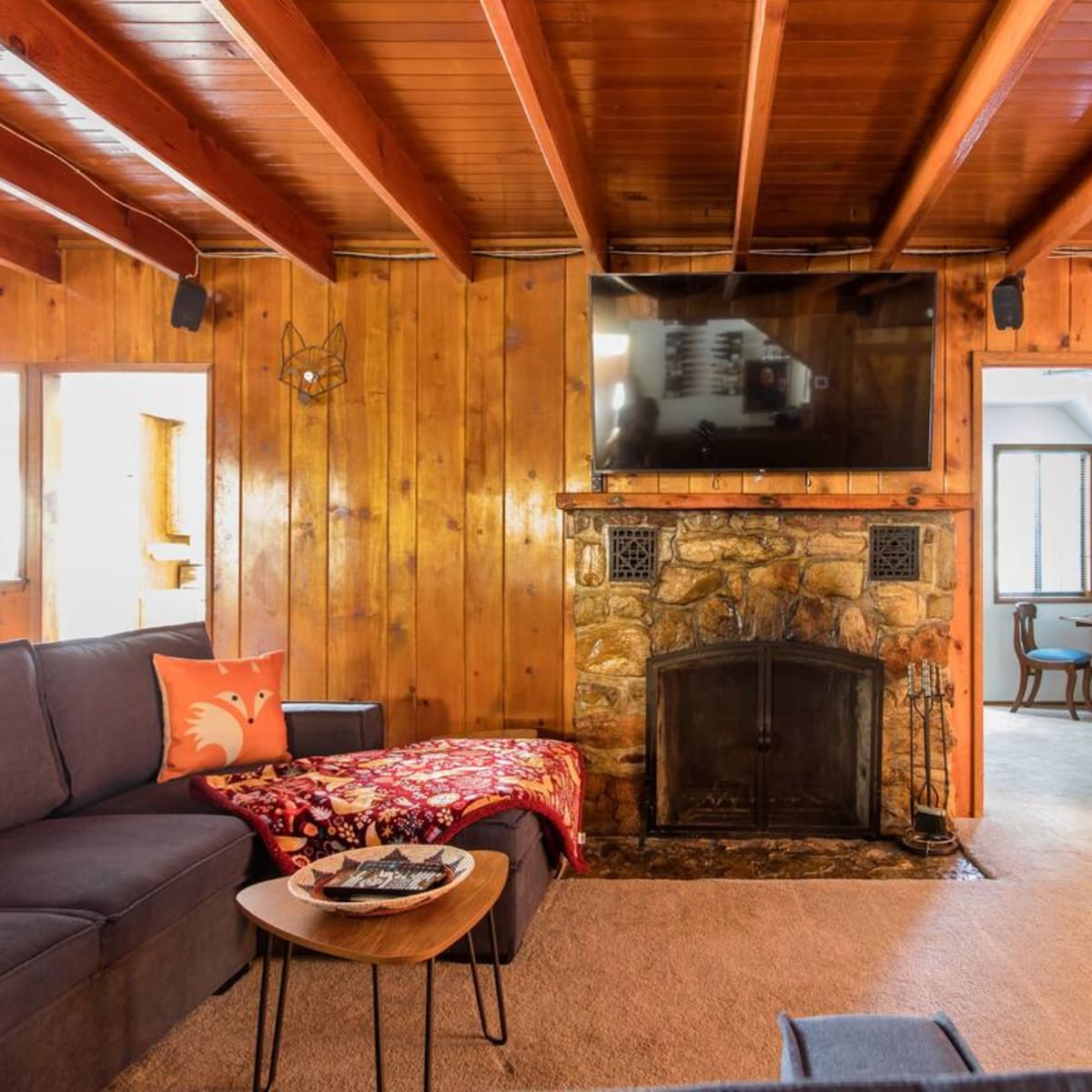 A cozy living room with wooden walls, a stone fireplace, a large TV mounted above it, and a sectional sofa with a decorative throw and a small coffee table.