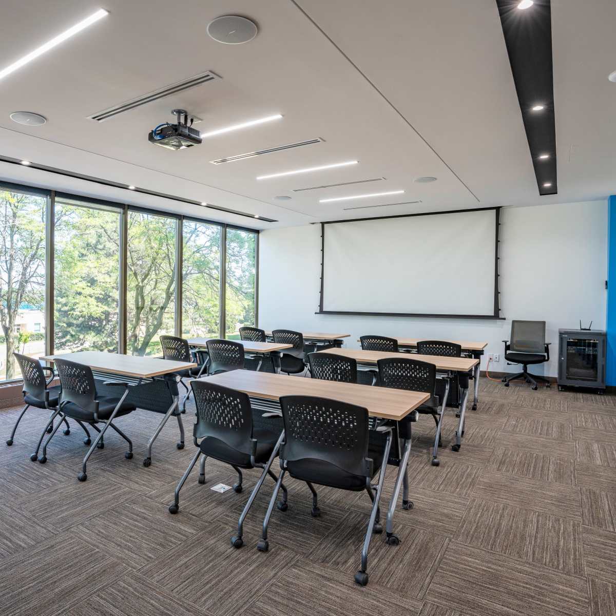 A modern conference room features a projector screen at the front, rows of tables and chairs, and large windows overlooking a green landscape.