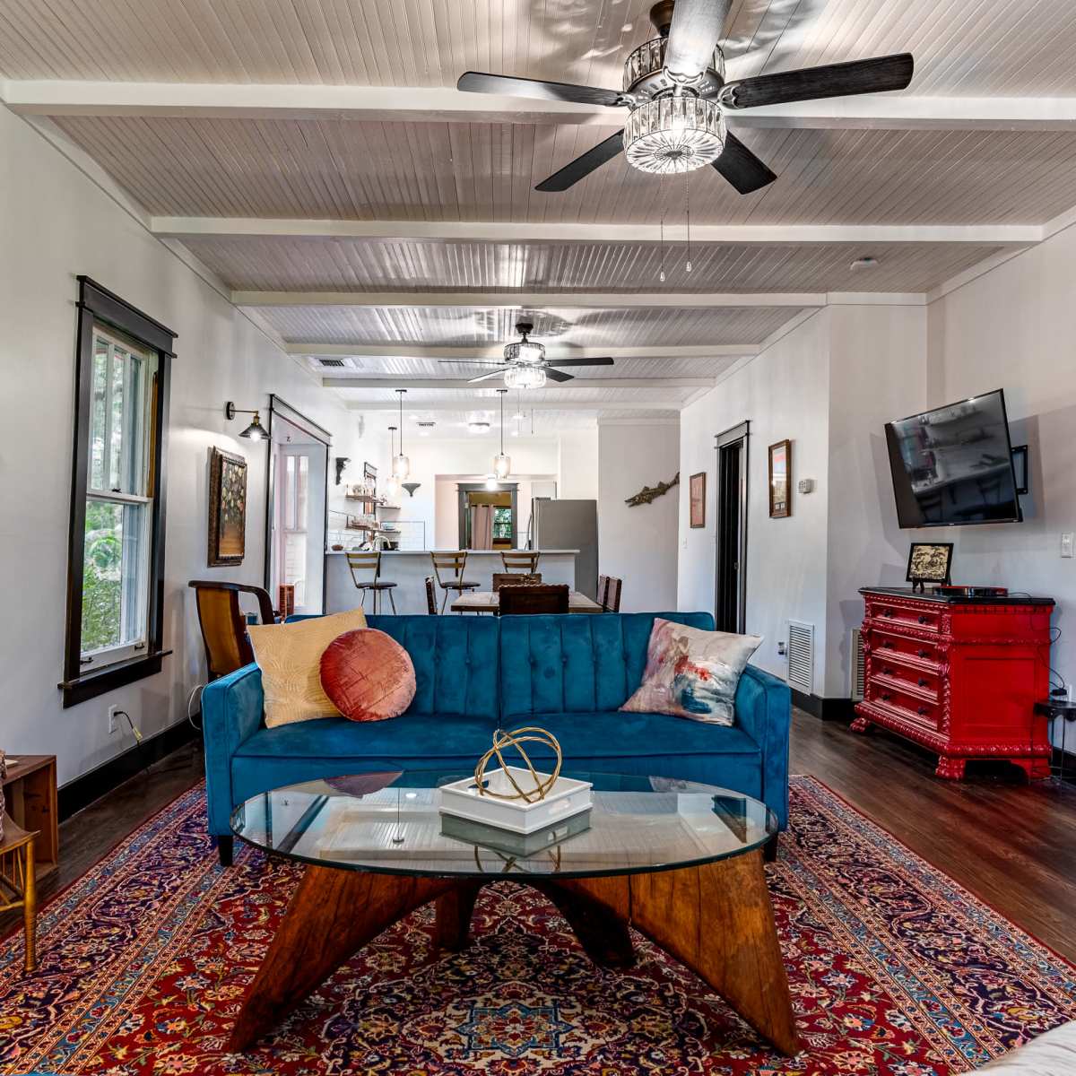 A living room featuring a blue velvet sofa, a glass-top coffee table, and a patterned rug, with an open kitchen visible in the background.