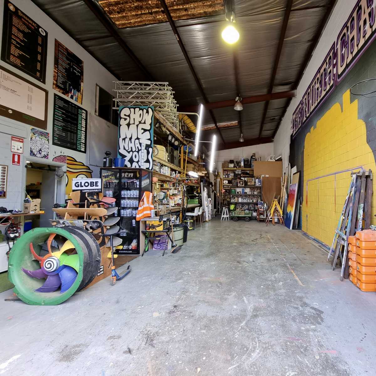 A large workshop space lined with shelves, tools, and materials, featuring a colorful mural on the wall and stacks of wood on one side.