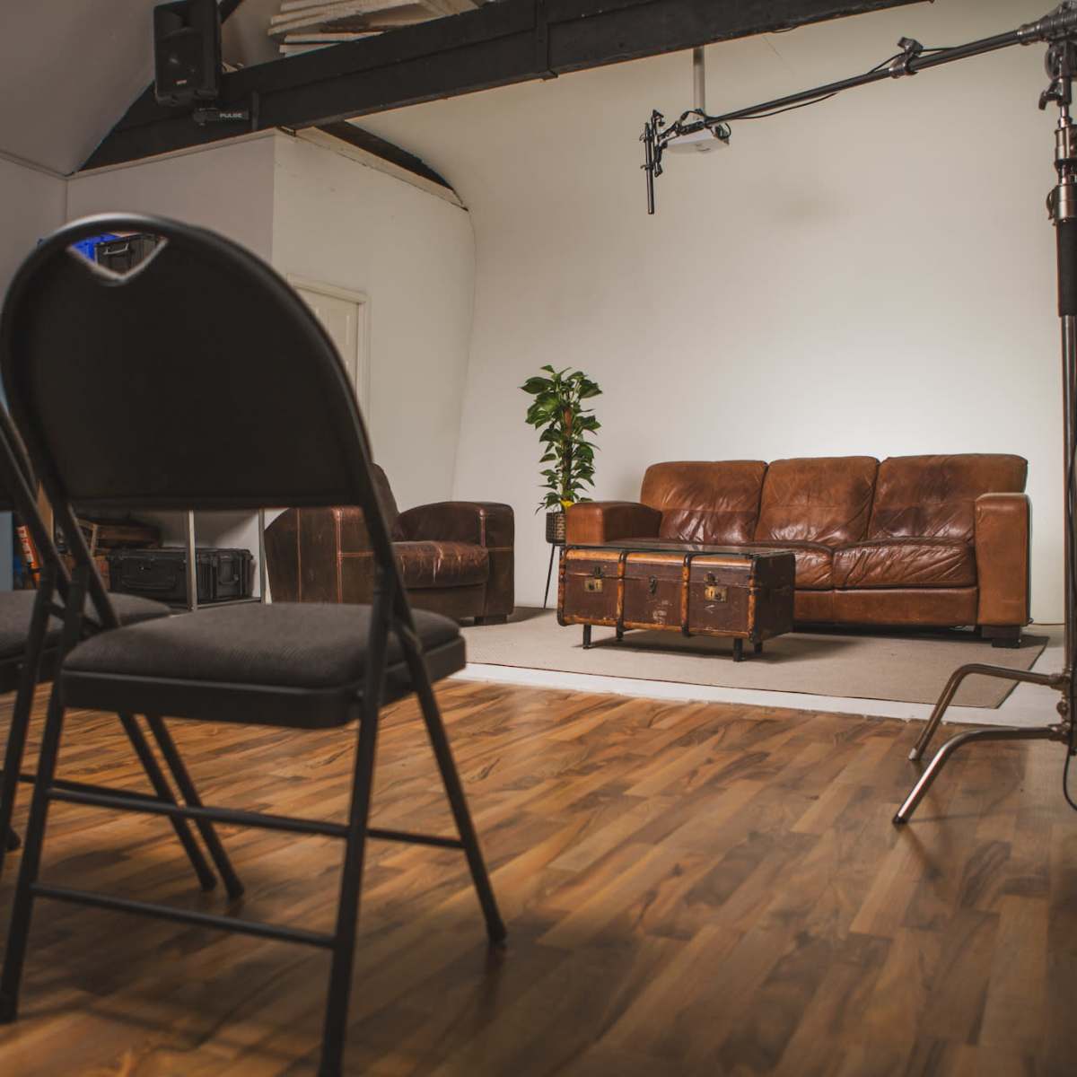 A room with a wooden floor, featuring rows of black folding chairs facing a brown leather sofa and a coffee table, with a microphone setup in front.
