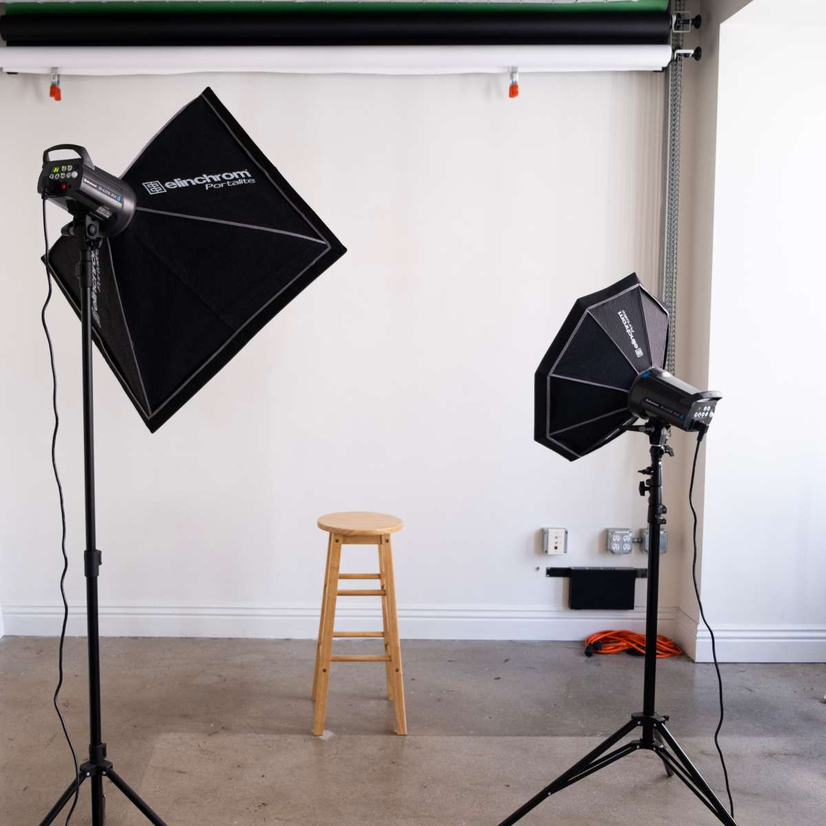 A photography studio setup with two light sources on tripods facing a wooden stool against a plain white wall.