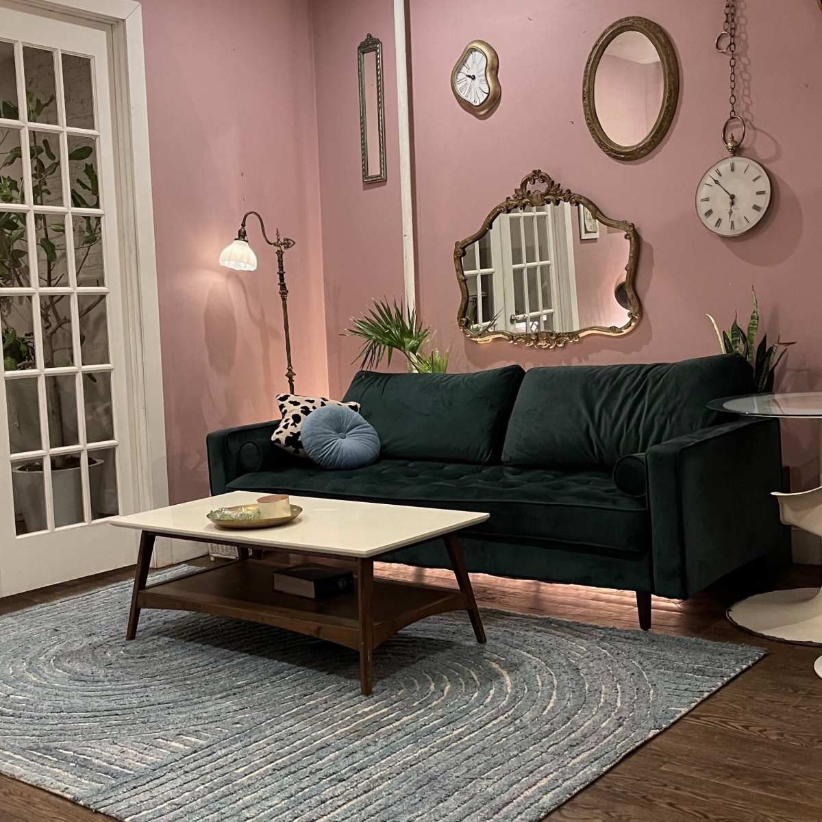 A cozy living room with a green velvet sofa, a round table with white chairs, and various decorative mirrors on pink walls.